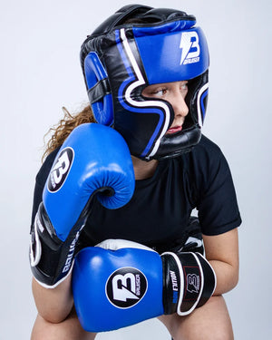 Casco Profesional Bruiser color azul y negro con rayas a los lados llevado por modelo mirando a un lado con brazos apoyados en rodillas y guantes azules. Logo en el centro. Diseñado con espuma densa y sistema de velcro ajustable. Diseño superior abierto. Imagen frontal.