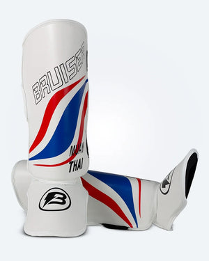 Espinilleras de muay thai Bruiser una de pie y otra estirada, blancas con diseño en azul y rojo inspirado en la bandera de Tailandia, logo Bruiser en la parte frontal y protección para espinilla y empeine para entrenamiento y kickboxing. Imagen frontal.
