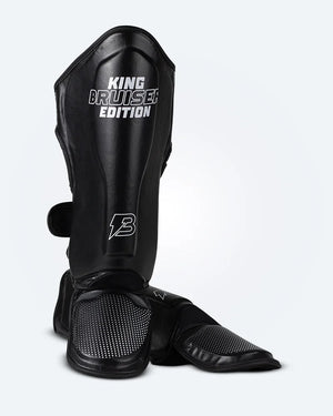 Espinilleras de Muay Thai "King Bruiser Edition" una de pie y otra estirada, color negro con detalles blancos. Logo Bruiser en la parte frontal inferior, hecha de piel sintética, protección para espinillas, tobillos y pies para Kickboxing, MMA y entrenamiento de contacto. Imagen frontal.