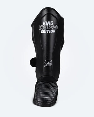 Espinillera de Muay Thai "King Bruiser Edition" color negro con detalles blancos. Logo Bruiser en la parte frontal inferior, hecha de piel sintética, protección para espinillas, tobillos y pies para Kickboxing, MMA y entrenamiento de contacto. Imagen frontal.