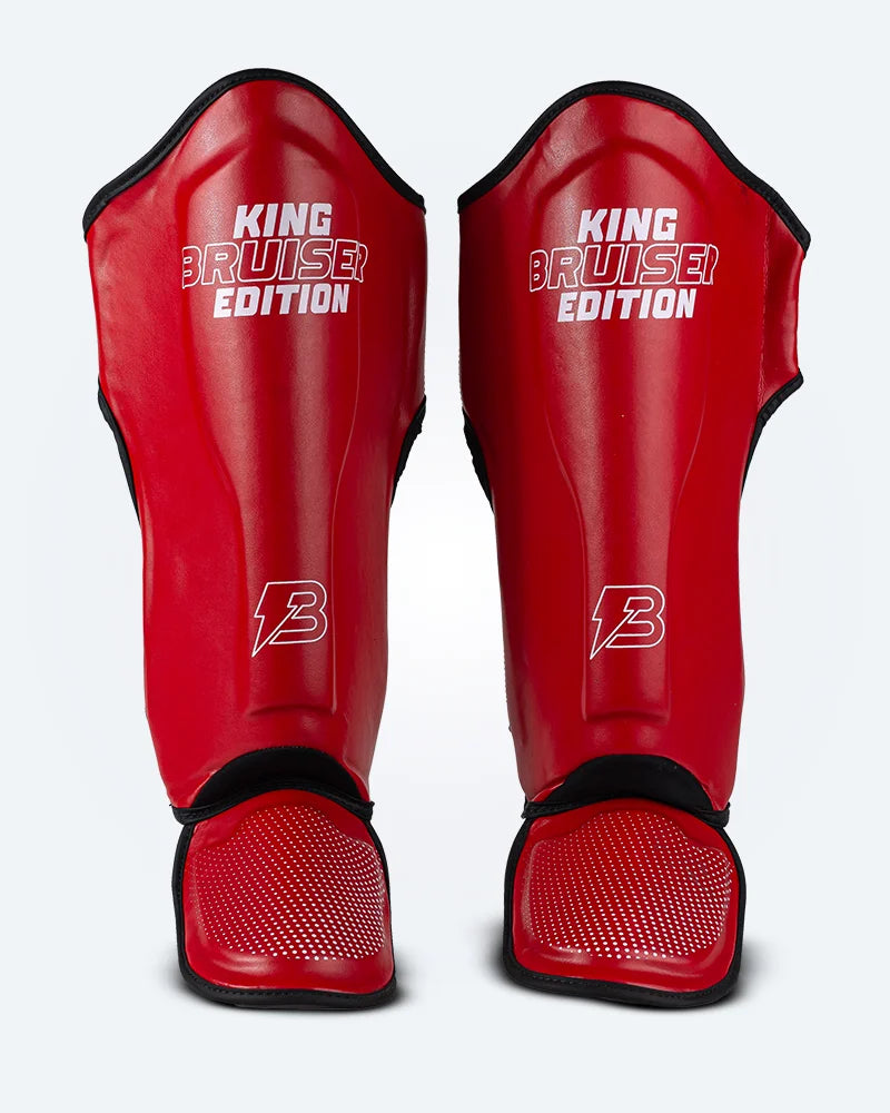 Espinilleras de Muay Thai "King Bruiser Edition" color rojo con detalles blancos. Logo Bruiser en la parte frontal inferior, hecha de piel sintética, protección para espinillas, tobillos y pies para Kickboxing, MMA y entrenamiento de contacto. Imagen frontal.