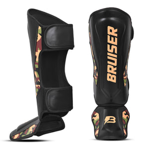 Espinilleras de Muay Thai "Bruiser CAMO", diseño de camuflaje combinado con negro y dorado, logo Bruiser en la parte frontal, hecha de piel sintética resistente, protección para espinillas, tobillos y pies. Para Kickboxing, MMA y entrenamiento de contacto. Imagen frontal.