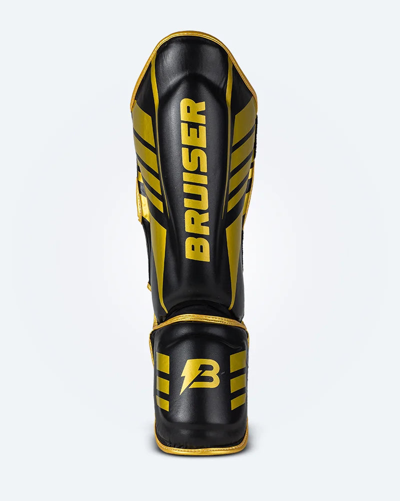 Espinillera de Muay Thai Bruiser color negro y dorado, logo Bruiser en la parte frontal y en empeine, hecha de piel sintética resistente, protección para espinillas, tobillos y pies. Para Kickboxing, MMA y entrenamiento de contacto. Imagen frontal.