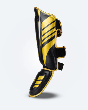 Perfil izquierdo espinillera de Muay Thai Bruiser color negro y dorado, logo Bruiser en la parte frontal y en empeine, hecha de piel sintética resistente, protección para espinillas, tobillos y pies, velcro reforzado. Para Kickboxing, MMA y entrenamiento de contacto.