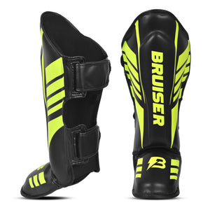 Espinilleras de kickboxing y muay thai negras con detalles verde neón y logo Bruiser en la parte frontal, diseño anatómico con protección para espinilla y empeine y cierre ajustable para entrenamiento y sparring.