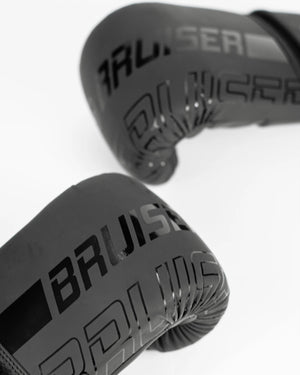 Par de guantillas Bruiser negras, logo brillante "Bruiser" en el centro en vertical. Hechas de polipiel resistente para entrenamientos frecuentes, golpeo en saco y manoplas. Imagen frontal.