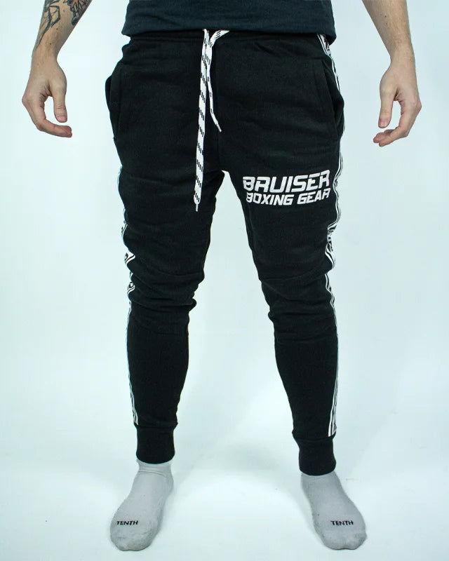 Pantalón de chándal negro con detalles blancos, estampado Bruiser en blanco en pierna izquierda, llevado por modelo de cintura con brazos estirados.