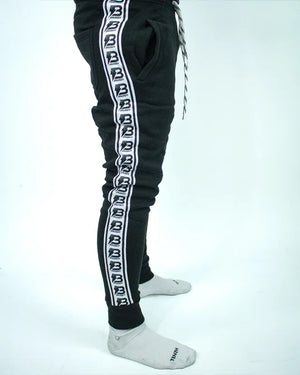 Perfil derecho de pantalón de chándal negro con banda lateral blanca y logos Bruiser repetidos, llevado por modelo de cintura con brazos estirados.