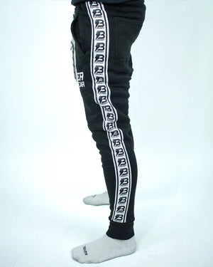 Perfil izquierdo de pantalón de chándal negro con banda lateral blanca y logos Bruiser repetidos, llevado por modelo de cintura con brazos estirados.