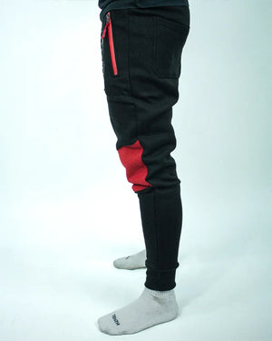 Perfil izquierdo de pantalón de chándal negro con detalles rojos, estampado Bruiser en rojo blanco y azul, llevado por modelo de cintura con brazos estirados.