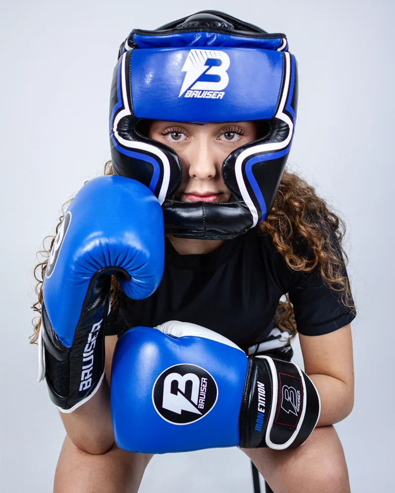 Casco Profesional Bruiser color azul y negro con rayas a los lados llevado por modelo con brazos apoyados en rodillas y guantes azules. Logo en el centro. Diseñado con espuma densa y sistema de velcro ajustable. Diseño superior abierto. Imagen frontal.
