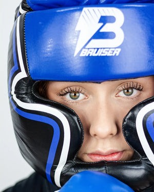 Casco Profesional Bruiser color azul y negro con rayas a los lados llevado por modelo mirando al frente. Logo en el centro. Diseñado con espuma densa y sistema de velcro ajustable. Diseño superior abierto. Imagen frontal.
