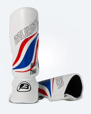 Espinilleras de muay thai Bruiser una de pie y otra estirada, blancas con diseño en azul y rojo inspirado en la bandera de Tailandia, logo Bruiser en la parte frontal y protección para espinilla y empeine para entrenamiento y kickboxing. Imagen frontal.