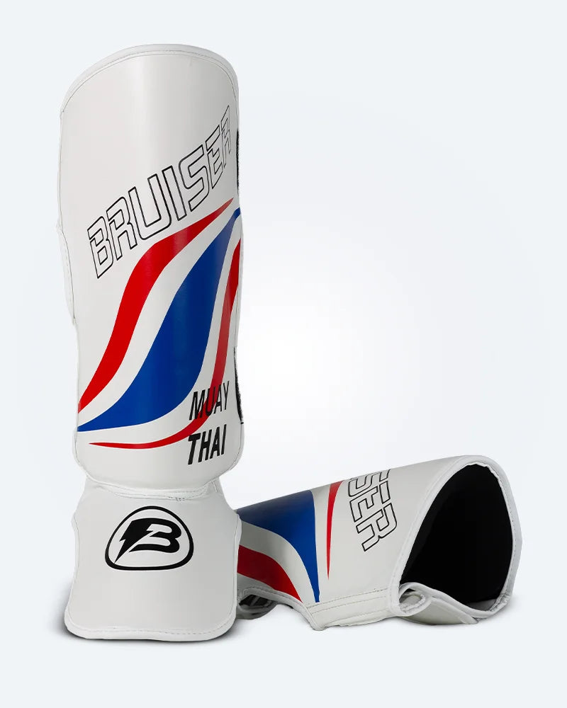 Espinilleras de muay thai Bruiser una de pie y otra estirada, blancas con diseño en azul y rojo inspirado en la bandera de Tailandia, logo Bruiser en la parte frontal y protección para espinilla y empeine para entrenamiento y kickboxing. Imagen frontal.