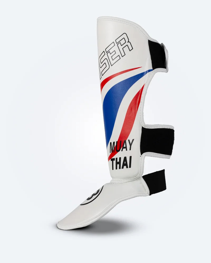 Perfil izquierdo de espinillera de kickboxing y muay thai, blanca con diseño en azul y rojo inspirado en la bandera de Tailandia, logo Bruiser en la parte frontal y protección para espinilla y empeine para entrenamiento y kickboxing. Ajuste de velcro seguro. 