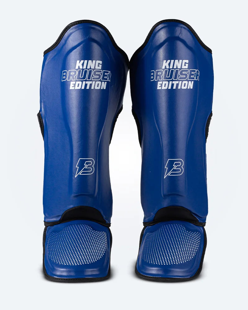 Espinilleras de Muay Thai "King Bruiser Edition" color azul con detalles blancos. Logo Bruiser en la parte frontal inferior, hecha de piel sintética, protección para espinillas, tobillos y pies para Kickboxing, MMA y entrenamiento de contacto. Imagen frontal. 