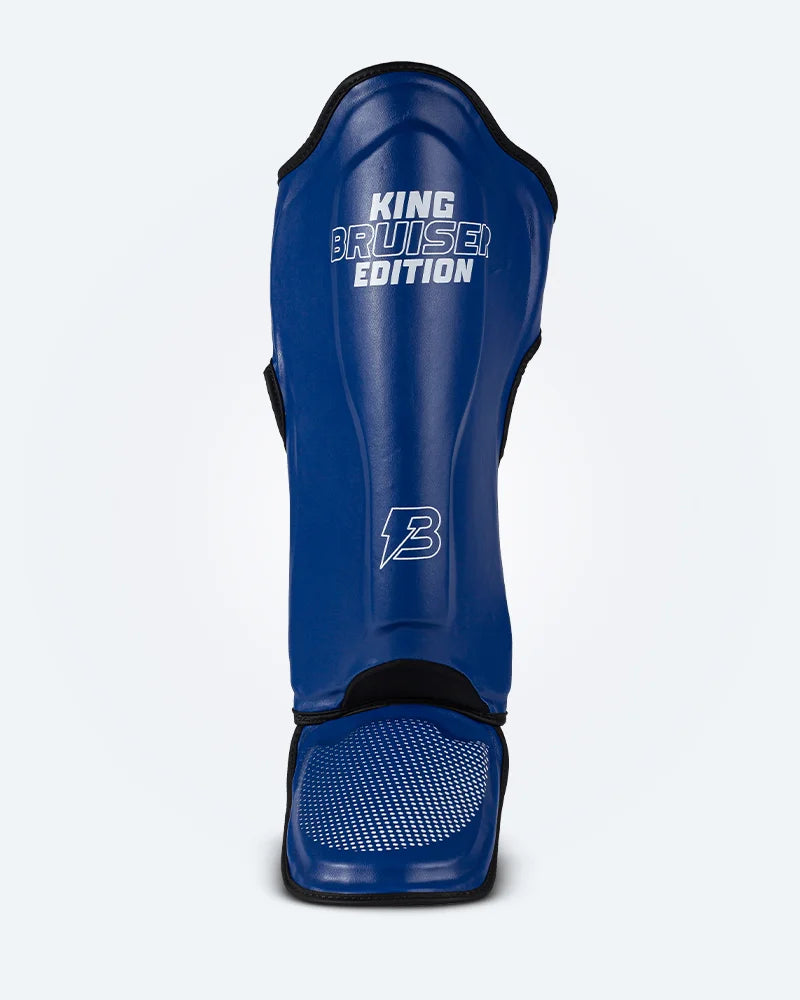 Espinillera de Muay Thai "King Bruiser Edition" color azul con detalles blancos. Logo Bruiser en la parte frontal inferior, hecha de piel sintética, protección para espinillas, tobillos y pies para Kickboxing, MMA y entrenamiento de contacto. Imagen frontal. 