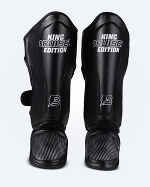 Espinilleras de Muay Thai "King Bruiser Edition" color negro con detalles blancos. Logo Bruiser en la parte frontal inferior, hecha de piel sintética, protección para espinillas, tobillos y pies para Kickboxing, MMA y entrenamiento de contacto. Imagen frontal. 