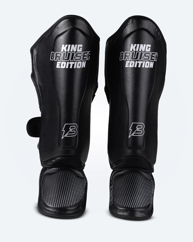 Espinilleras de Muay Thai "King Bruiser Edition" color negro con detalles blancos. Logo Bruiser en la parte frontal inferior, hecha de piel sintética, protección para espinillas, tobillos y pies para Kickboxing, MMA y entrenamiento de contacto. Imagen frontal. 