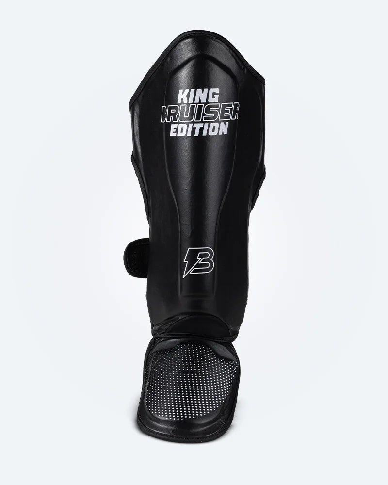 Espinillera de Muay Thai "King Bruiser Edition" color negro con detalles blancos. Logo Bruiser en la parte frontal inferior, hecha de piel sintética, protección para espinillas, tobillos y pies para Kickboxing, MMA y entrenamiento de contacto. Imagen frontal. 