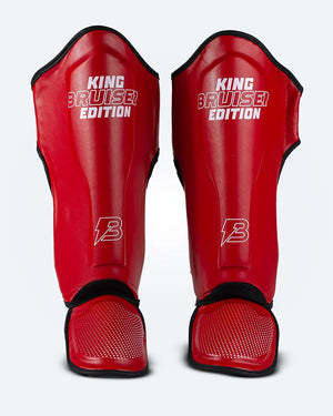 Espinilleras de Muay Thai "King Bruiser Edition" color rojo con detalles blancos. Logo Bruiser en la parte frontal inferior, hecha de piel sintética, protección para espinillas, tobillos y pies para Kickboxing, MMA y entrenamiento de contacto. Imagen frontal. 