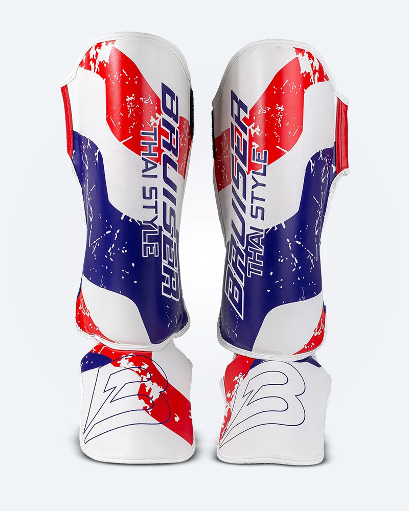 Espinilleras de Muay Thai Bruiser blancas con diseño en azul y rojo inspirado en la bandera de Tailandia, logo Bruiser en la parte frontal, hecha de piel sintética, protección para espinilla y empeine para entrenamiento y kickboxing. Imagen frontal.