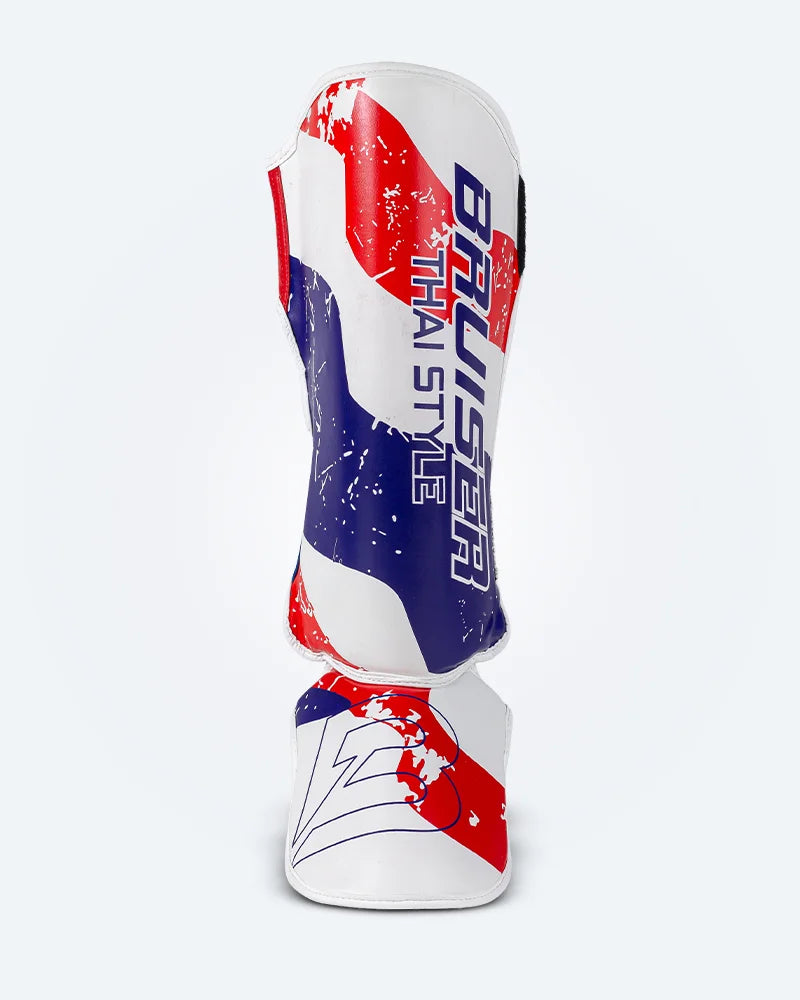 Espinillera de Muay Thai Bruiser blanca con diseño en azul y rojo inspirado en la bandera de Tailandia, logo Bruiser en la parte lateral, hecha de piel sintética, protección para espinilla y empeine para entrenamiento y kickboxing. Imagen frontal.