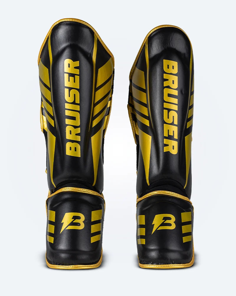 Espinilleras de Muay Thai Bruiser color negro y dorado, logo Bruiser en la parte frontal y en empeine, hecha de piel sintética resistente, protección para espinillas, tobillos y pies. Para Kickboxing, MMA y entrenamiento de contacto. Imagen frontal. 