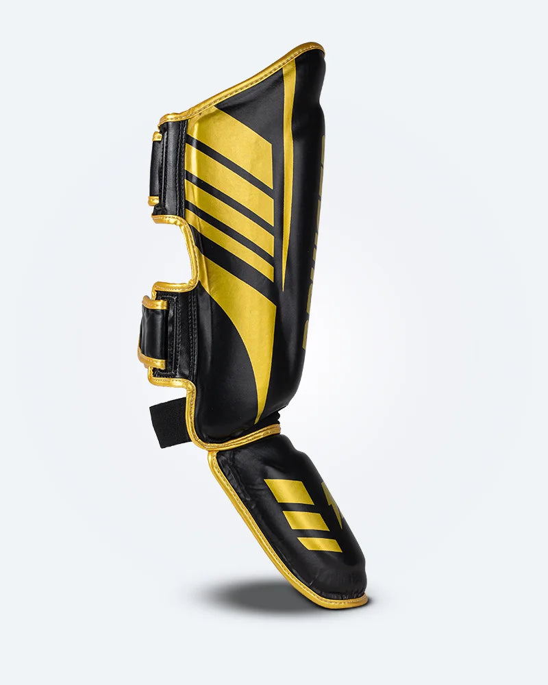 Perfil derecho espinillera de Muay Thai Bruiser color negro y dorado, logo Bruiser en la parte frontal y en empeine, hecha de piel sintética resistente, protección para espinillas, tobillos y pies, velcro reforzado. Para Kickboxing, MMA y entrenamiento de contacto. 