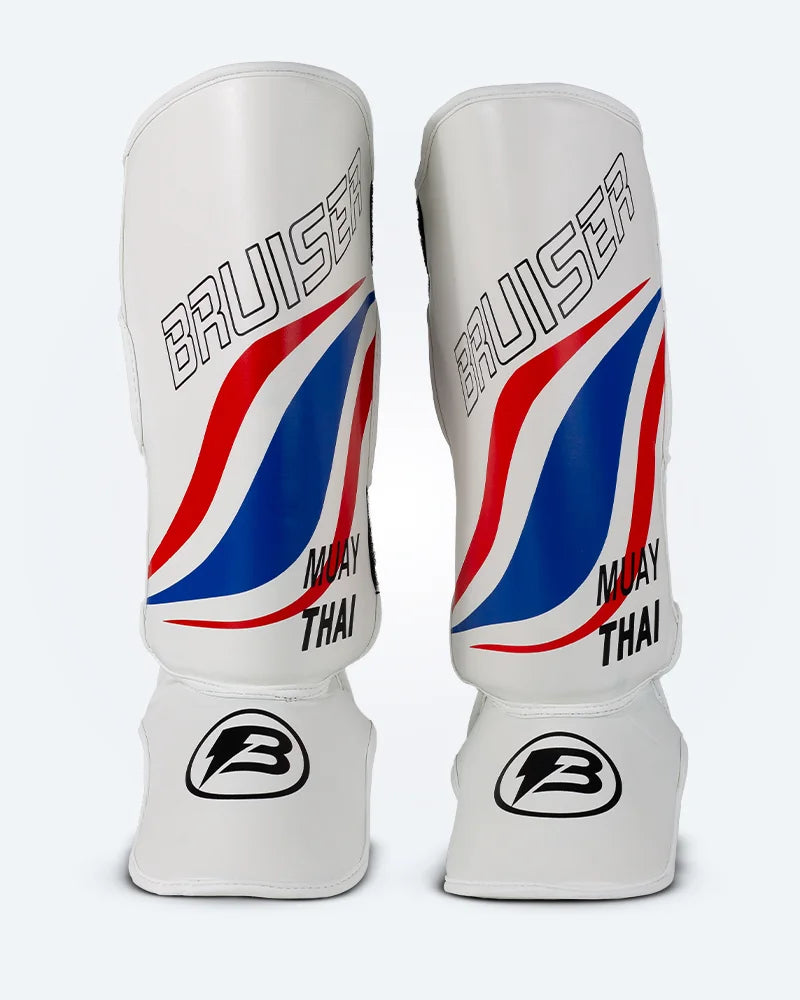 Espinilleras de muay thai Bruiser blancas con diseño en azul y rojo inspirado en la bandera de Tailandia, logo Bruiser en la parte frontal y protección para espinilla y empeine para entrenamiento y kickboxing. Imagen frontal.