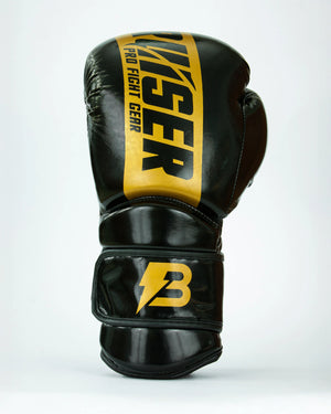 Guantes de boxeo Bruiser color negro y dorado. Logo de la marca en el centro en vertical. Hecho de polipiel resistente con acolchado interior. Cierre de velcro seguro. Para entrenamiento en saco y sparring controlado. Imagen frontal. 