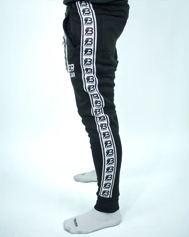 Perfil izquierdo de pantalón de chándal negro con banda lateral blanca y logos Bruiser repetidos, llevado por modelo de cintura con brazos estirados. 