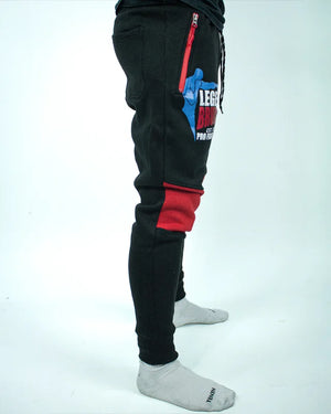 Perfil derecho de pantalón de chándal negro con detalles rojos, estampado Bruiser en rojo blanco y azul, llevado por modelo de cintura con brazos estirados. 