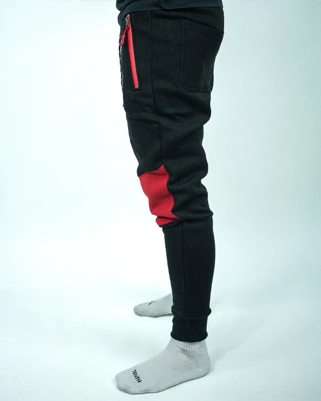 Perfil izquierdo de pantalón de chándal negro con detalles rojos, estampado Bruiser en rojo blanco y azul, llevado por modelo de cintura con brazos estirados. 