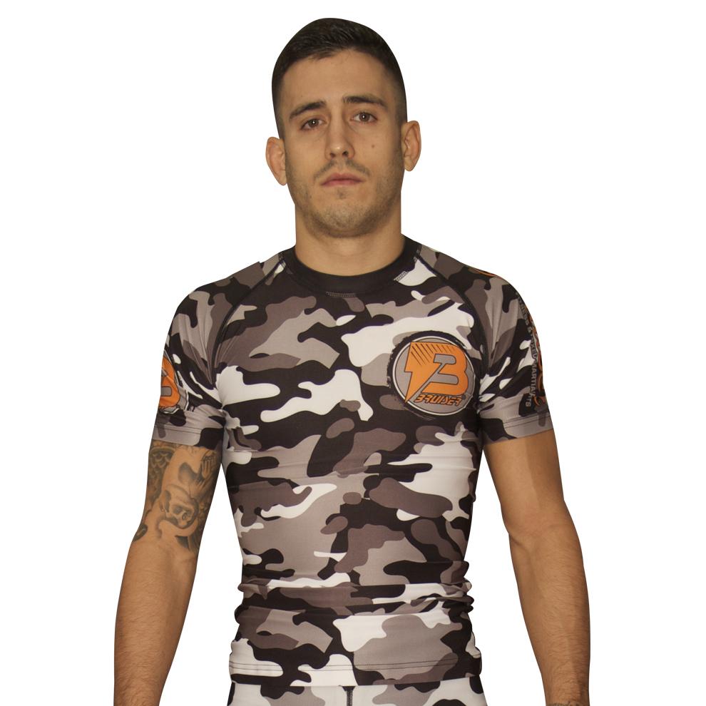 Camisera deportiva Rash Guard ceñida de la marca "Bruiser" diseño camuflaje con logo en naranja. Llevada por modelo con brazos estirados. Transpirable con diseño sublimado. Imagen frontal.  